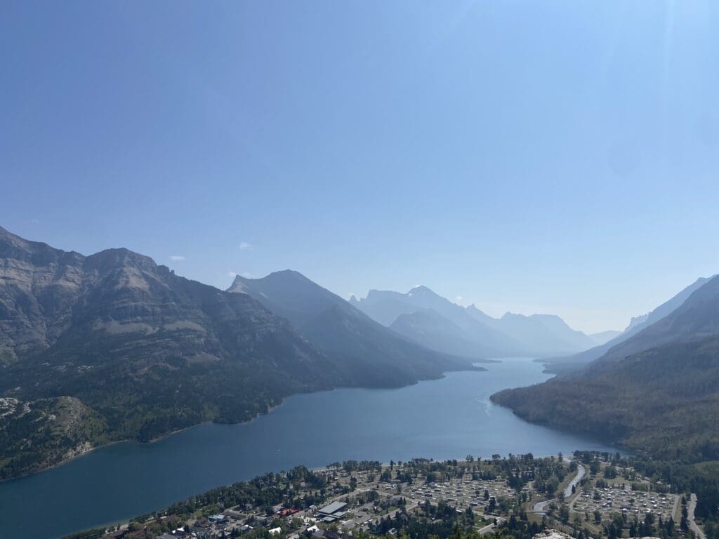 A serene landscape of Waterton Lake in Waterton Lakes National Park, Alberta surrounded by mountains under a clear sky. In the foreground, there is a small town with scattered buildings.
