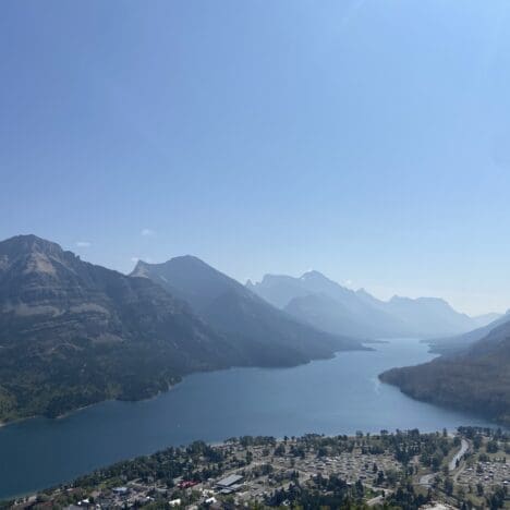A serene landscape of Waterton Lake in Waterton Lakes National Park, Alberta surrounded by mountains under a clear sky. In the foreground, there is a small town with scattered buildings.