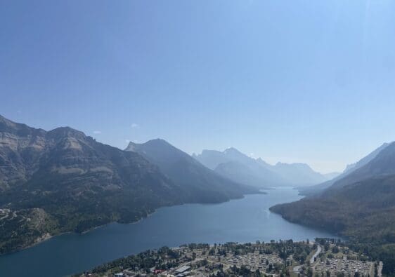 A serene landscape of Waterton Lake in Waterton Lakes National Park, Alberta surrounded by mountains under a clear sky. In the foreground, there is a small town with scattered buildings.
