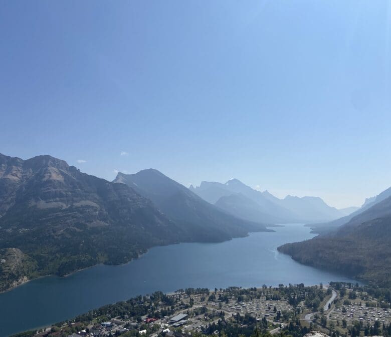 A serene landscape of Waterton Lake in Waterton Lakes National Park, Alberta surrounded by mountains under a clear sky. In the foreground, there is a small town with scattered buildings.
