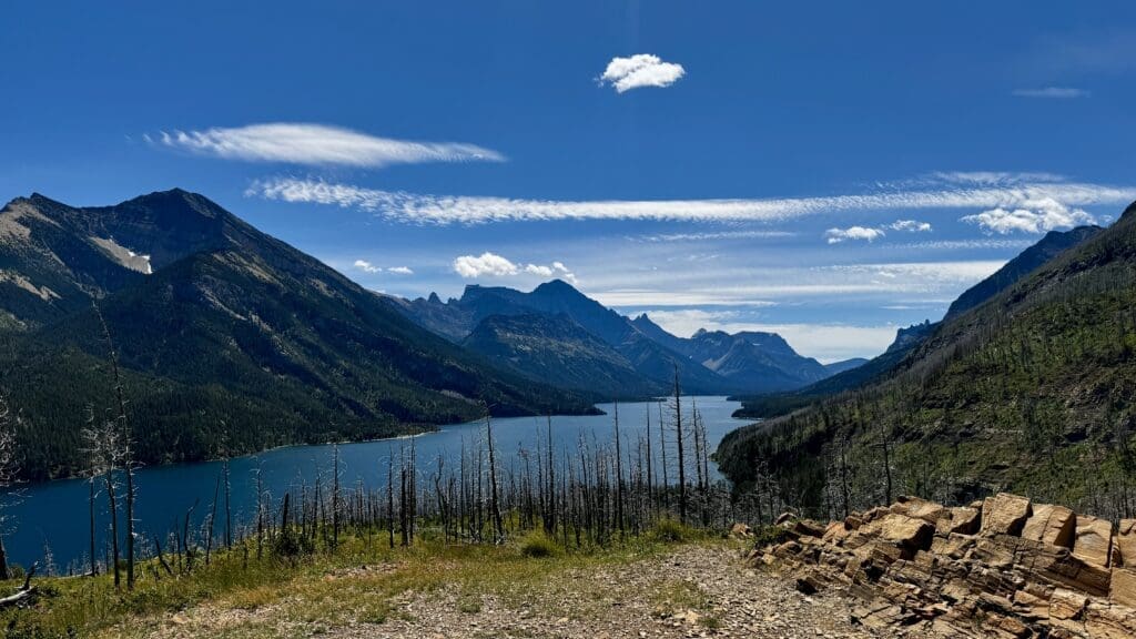 Mountainous landscape with the serene blue Waterton Lake in Waterton Lakes National Park, Alberta surrounded by rugged peaks under a vibrant blue sky. Bare trees and rocky foreground add depth. Calm and majestic.