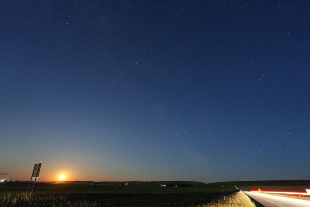 Night sky with stars in Pincher Creek, Alberta above a rural landscape, where the moon rises on the horizon. A road with blurred car lights adds motion to the serene scene.