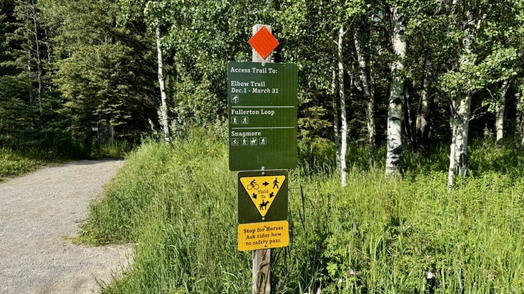 Trail sign surrounded by lush green forest. It displays directions for Elbow Trail, Fullerton Loop, and Snagmore in Kananaskis, Alberta. Cautionary signs for cyclists and pedestrians.