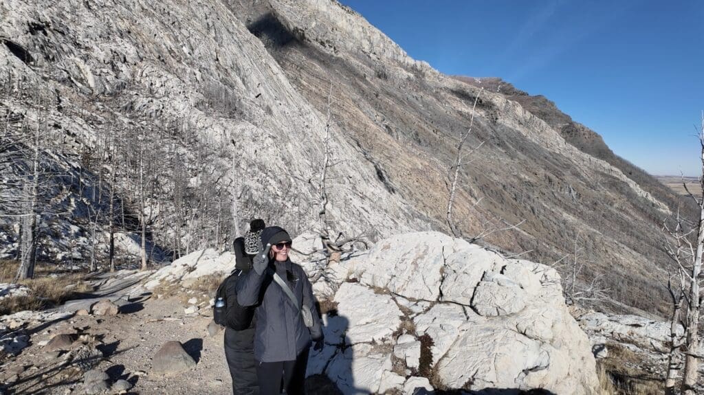 A person in a winter jacket and beanie and another person in windproof jacket stands on a rocky mountain Bear's Hump Trail in Waterton Lakes National Park, Alberta. They smile, flashing a peace sign. Barren trees and rugged cliffs surround them under a clear blue sky.