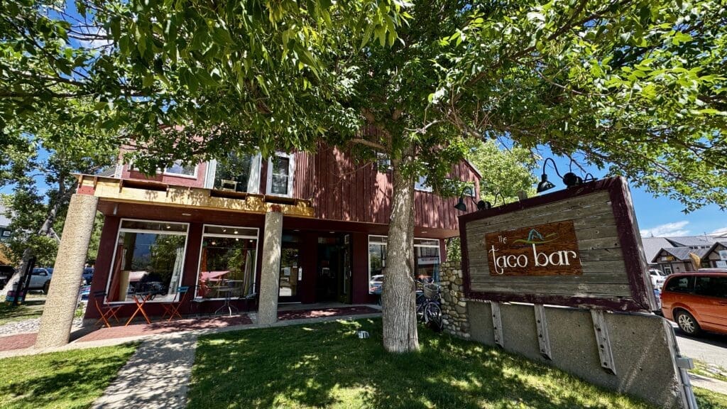 Red building in Waterton Townsite, Alberta with a sign reading "The Taco Bar" featuring a shaded patio. Green lawn, leafy trees, and a bright sunny atmosphere convey a welcoming vibe.
