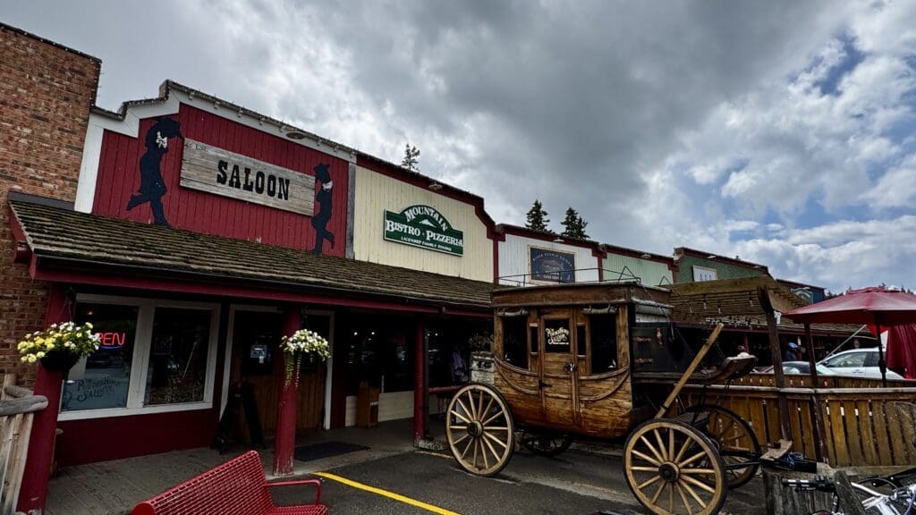 Western-style storefronts in Bragg Creek, Alberta, including a saloon and bistro, under cloudy skies. A vintage wooden carriage is parked in front, creating a nostalgic atmosphere.
