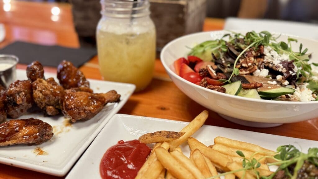 A plate of glazed chicken wings with dipping sauce, crispy fries with ketchup, a vibrant salad with mixed greens, and a mason jar of lemonade on a wooden table in Thirsty Bear Kitchen + Bar in Waterton Townsite, Alberta.