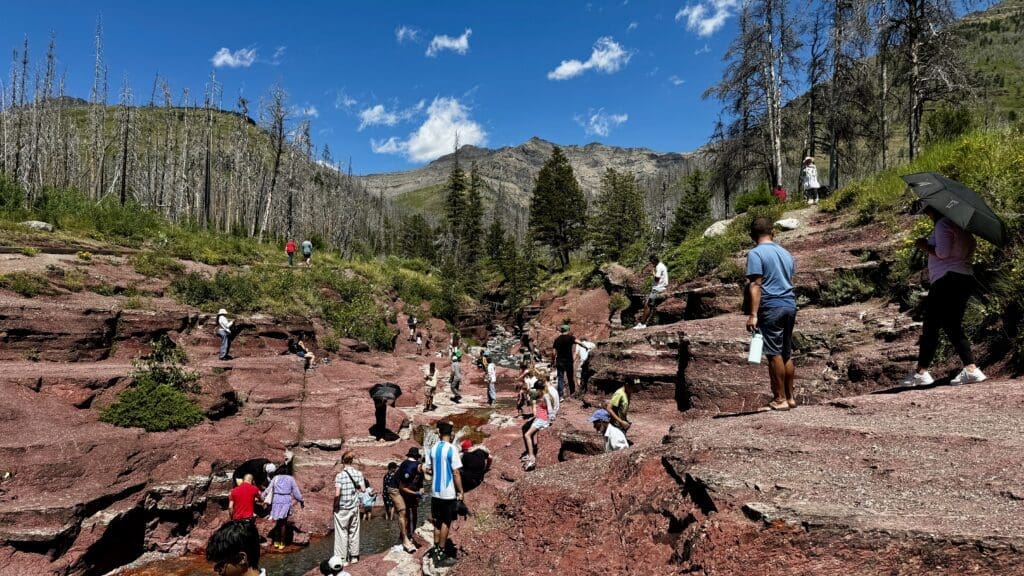 People explore Red Rock Canyon in Waterton Lakes National Park, Alberta with sparse trees, under a clear blue sky. The mood is adventurous and lively.