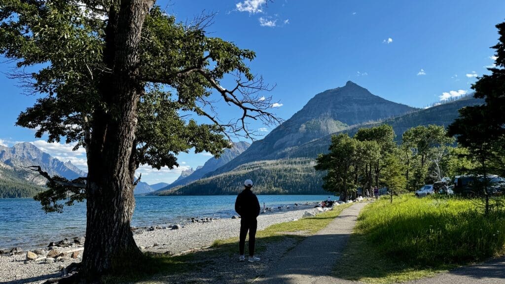 A person in a white cap stands near the tranquil Waterton Lake in Waterton Lakes National Park, Alberta, surrounded by lush greenery and mountains under a clear blue sky. The scene is peaceful and scenic.