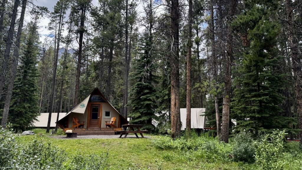 A-frame cabin with a wooden porch nestled among tall pine trees in a lush forest in Two Jack Lake in Banff National Park, Alberta. Nearby, there is a picnic table and a fire pit, creating a serene, rustic setting.