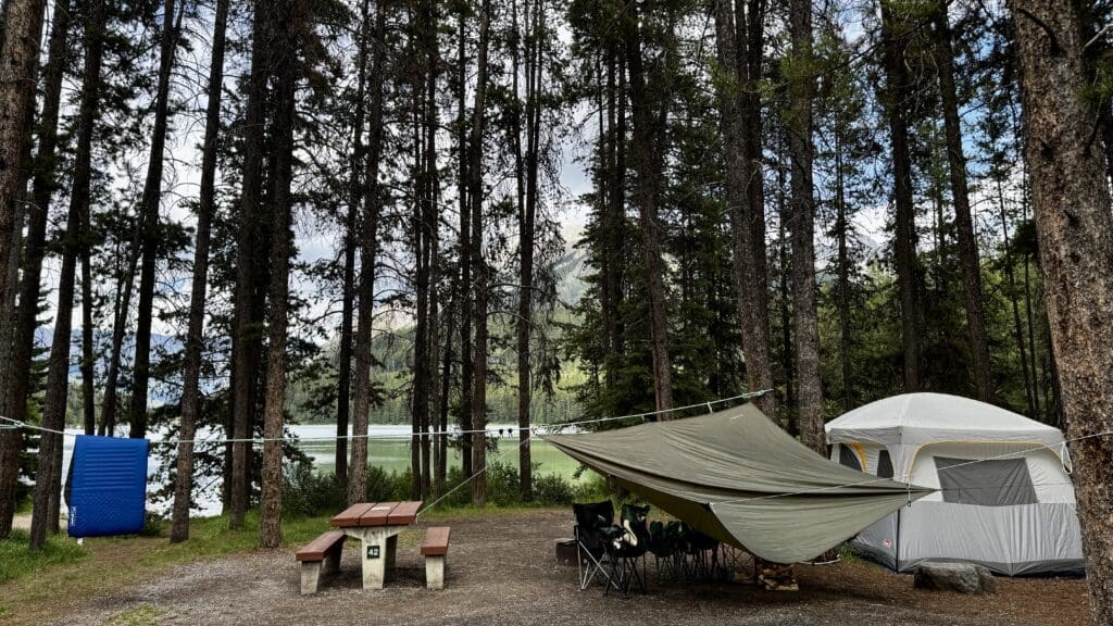 Camping scene in a forest by Two Jack Lake in Banff National Park, Alberta. A tent and tarp shelter with chairs are surrounded by tall trees. A picnic table and blue mat complete the serene setup.