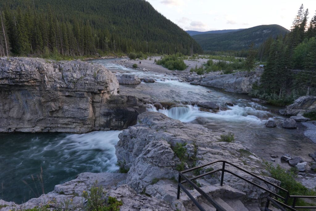 A tranquil Elbow River in Kananaskis, Alberta flows over rocky cascades surrounded by lush evergreen forests and mountains. The scene, viewed from atop stairs, evokes peacefulness.