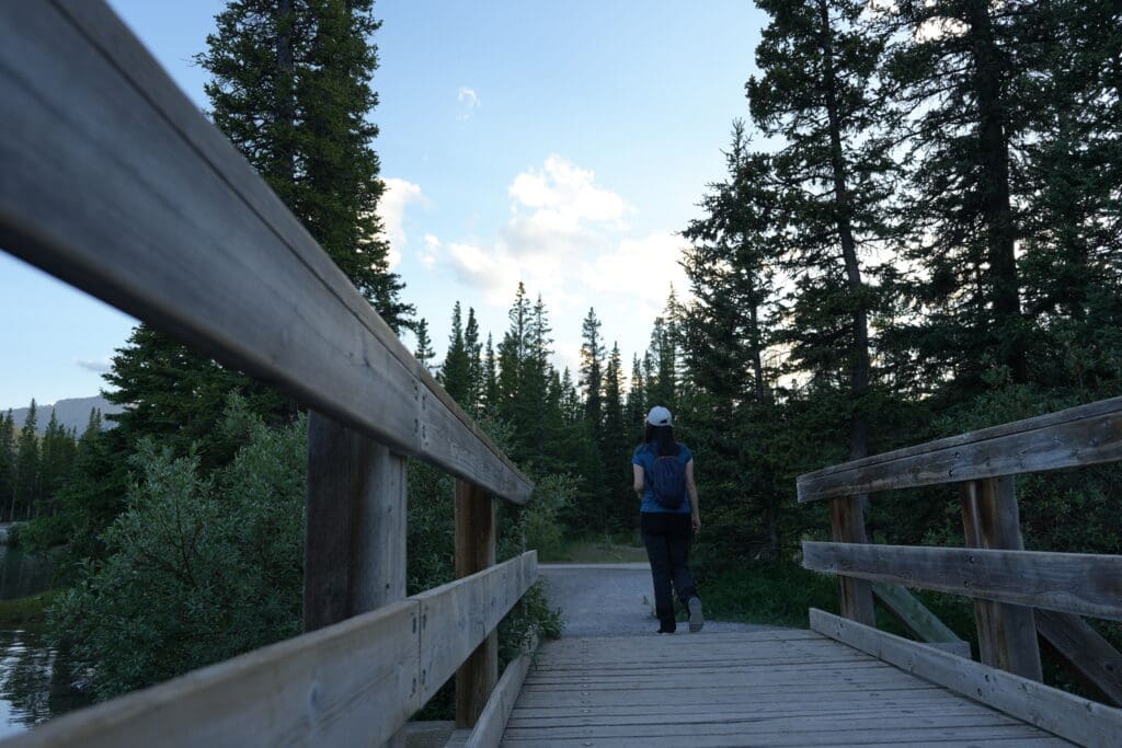 A person with a backpack walks across a wooden bridge along Forgetmenot Pond in Kananaskis, Alberta toward a pine forest under a clear sky. The serene scene evokes a sense of adventure.