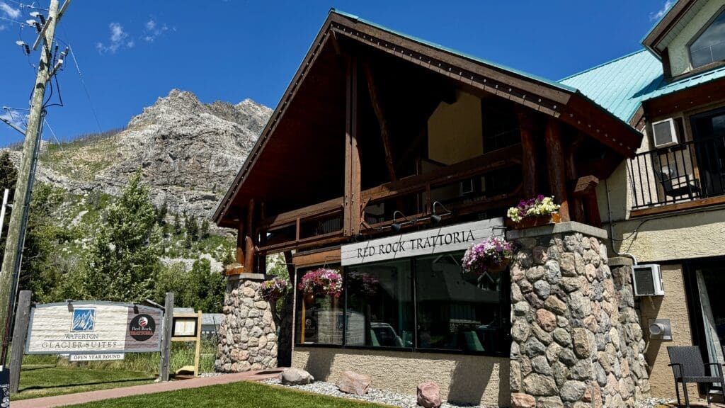 Charming mountain restaurant, "Red Rock Trattoria," in Waterton Glacier Suites in Waterton Townsite, Alberta with stone and wood facade. Hanging flower baskets add vibrant color under a clear blue sky.