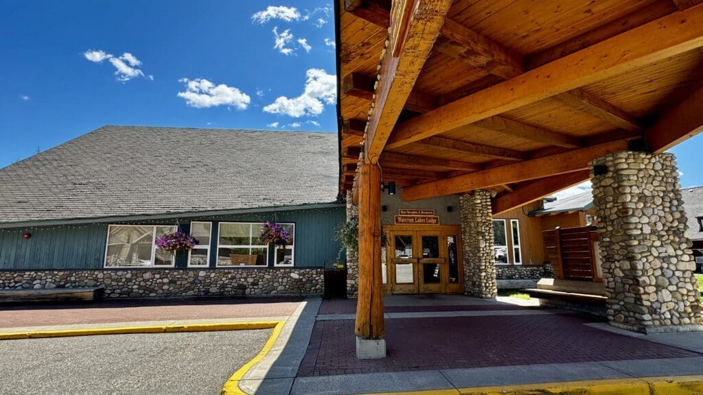Rustic lodge entrance of Waterton Lakes Lodge Resort in Waterton Townsite, Alberta with wooden beams and stone pillars under a clear blue sky. Purple flowers hang by windows, evoking a welcoming, serene atmosphere.