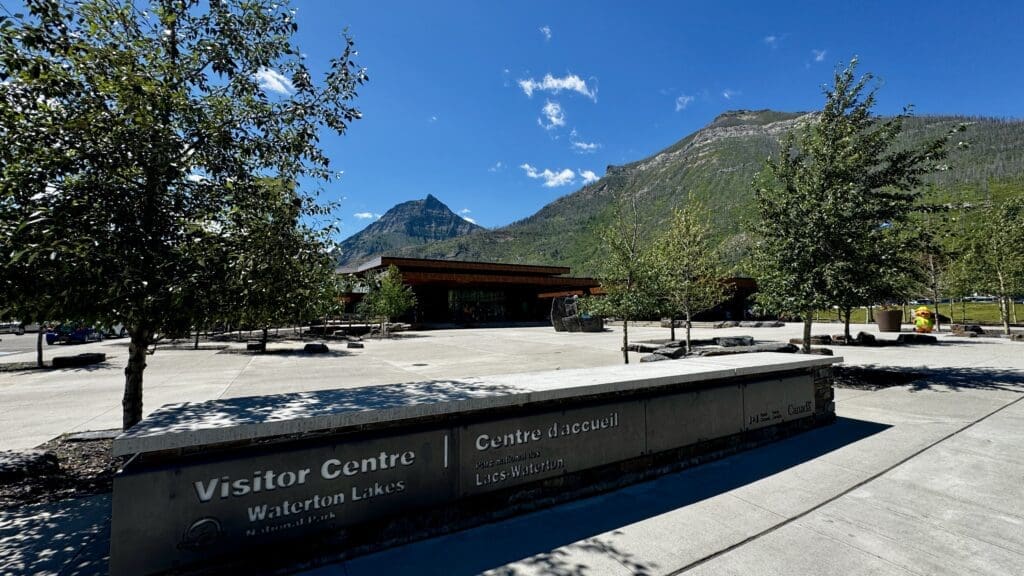 Visitor Centre at Waterton Lakes National Park, Alberta with a modern building, surrounded by trees and mountains under a clear blue sky.