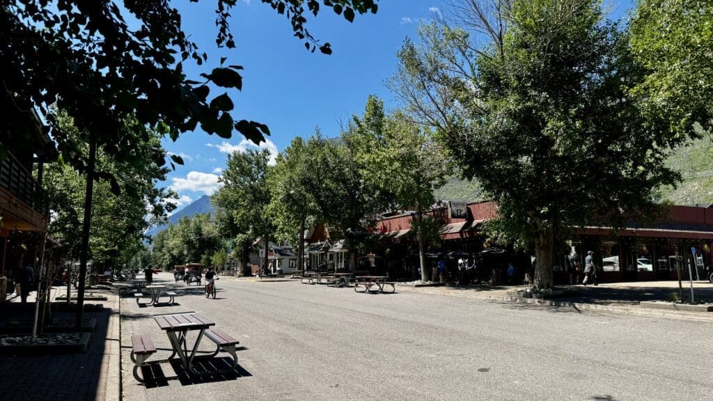 Sunny street scene in Waterton Townsite in Waterton Lakes National Park, Alberta with trees lining a quiet road, cyclists riding by, picnic tables on the sidewalk, and an inviting row of shops under a clear blue sky.