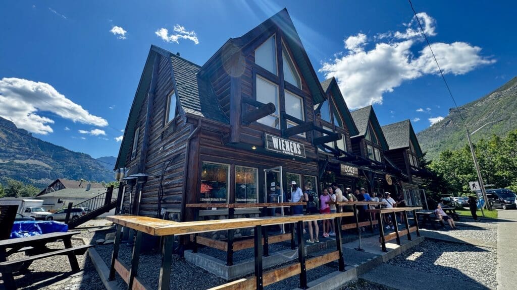 Alpine-style wooden building under a blue sky, with a "Wieners" sign in Waterton Townsite, Alberta. A queue of people waits outside, evoking a lively and inviting atmosphere.